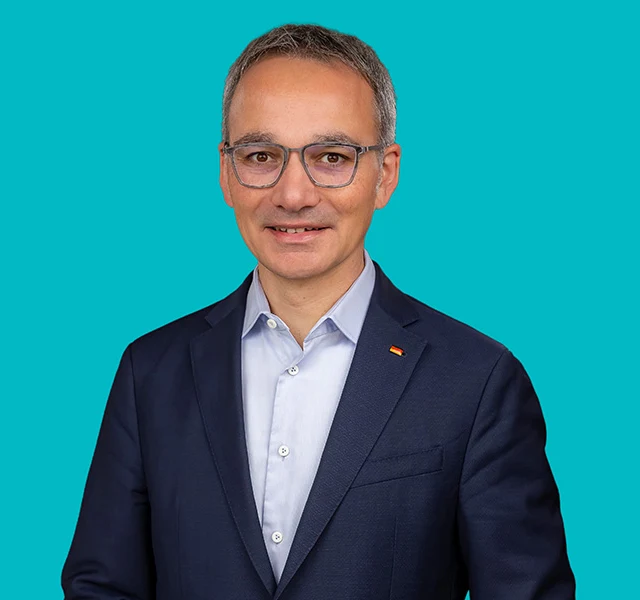 Wilfried Oellers im dunkelblauen Sakko vor türkisem Hintergrund