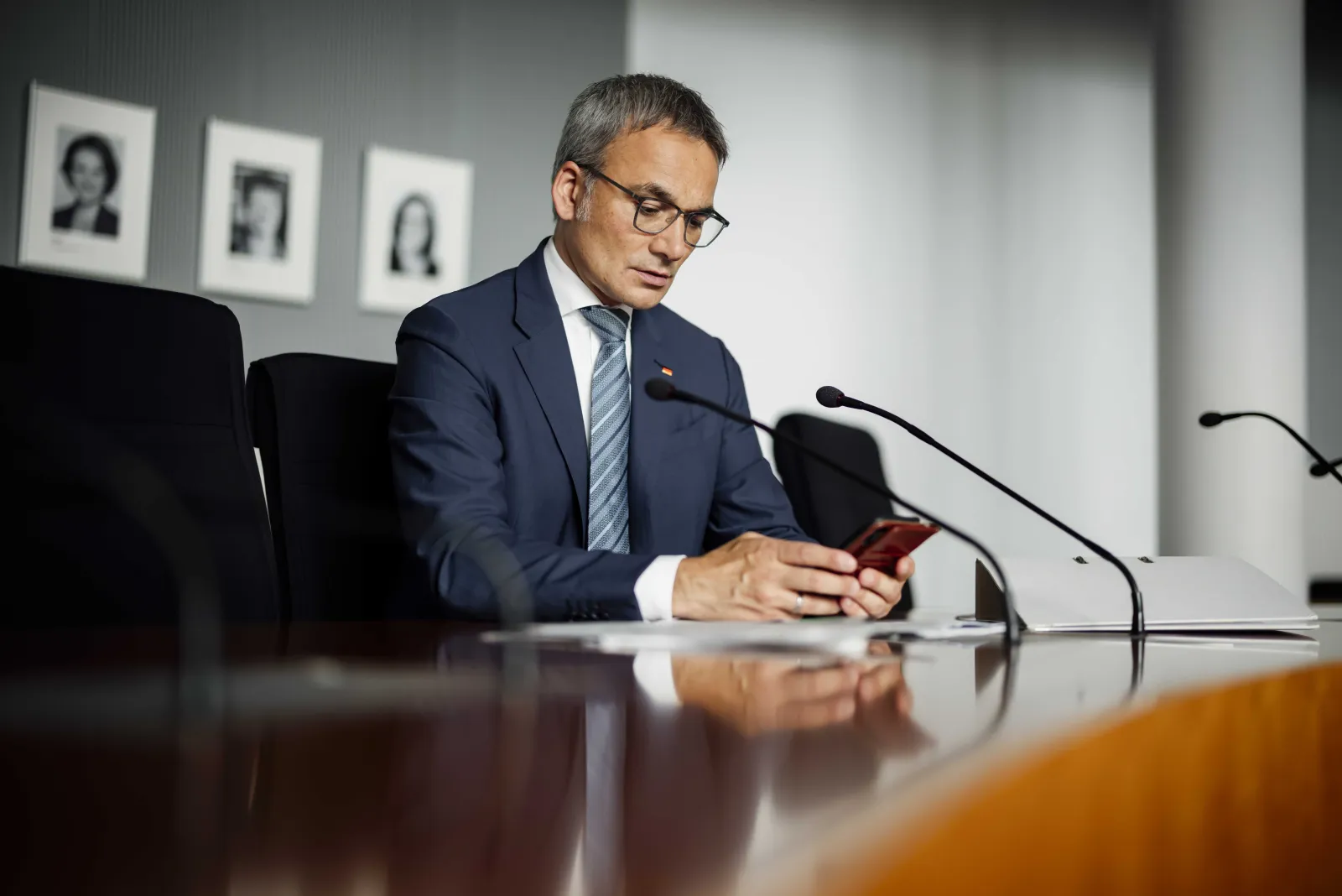 Wilfried Oellers an einem Sitzungstisch mit Mikrofonen, den Blick auf sein Smartphone gerichtet
